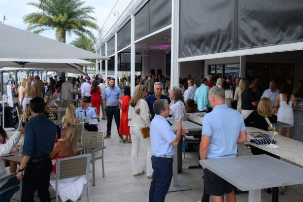 A large group of people socializing at an outdoor restaurant or bar with open walls, palm trees, and relaxed, summery attire. Dining tables and umbrellas are visible, creating a lively, upscale atmosphere. South Florida Business & Wealth