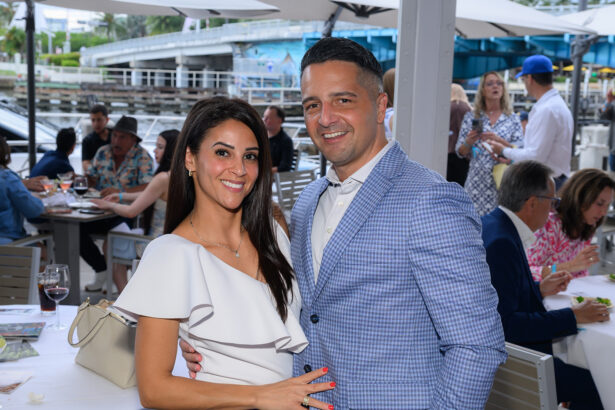 A smiling couple poses together at an outdoor waterfront restaurant, dressed in semi-formal attire. Other guests are dining and socializing in the background under umbrellas. South Florida Business & Wealth