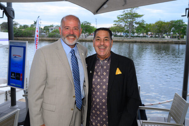 Two men in suits smile and pose together by a waterfront under a patio umbrella. Trees and water are in the background, and one man wears a tan suit while the other wears a black patterned suit. South Florida Business & Wealth