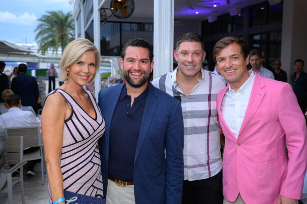 Four people pose and smile at an outdoor event; a woman in a pink striped dress stands beside three men in jackets and shirts, with palm trees and a patio area visible in the background. South Florida Business & Wealth