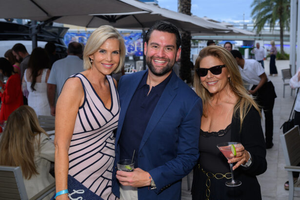 Three people stand outdoors at a social event, smiling at the camera. The woman on the left wears a light striped dress, the man in the middle wears a blue blazer, and the woman on the right holds a cocktail. South Florida Business & Wealth