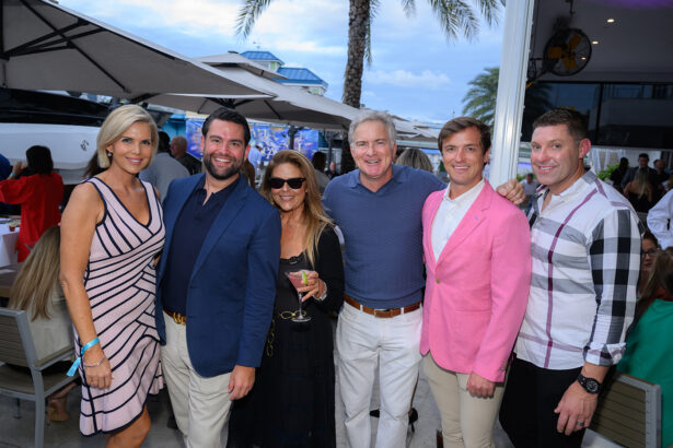 Six adults dressed in semi-formal attire pose and smile together at an outdoor event by palm trees and yachts. Some hold drinks and the group appears happy and relaxed under a partly cloudy sky. South Florida Business & Wealth