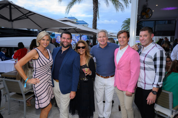 Six people dressed in semi-formal attire smile for a group photo at an outdoor event by the water, with palm trees, tables, and umbrellas in the background. South Florida Business & Wealth