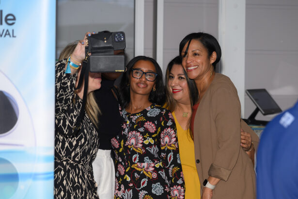 Four women stand close together and smile as one of them takes a selfie with a camera. They are indoors and appear to be at an event or gathering. South Florida Business & Wealth