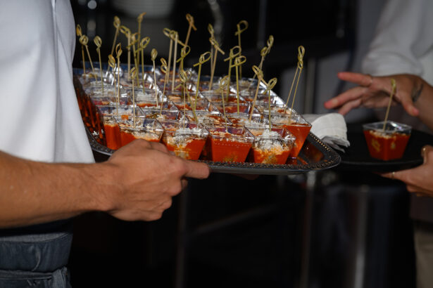 A server holds a tray of small appetizer cups garnished with cheese and skewers, while another person reaches to take one. The setting appears to be a catered event or party. South Florida Business & Wealth