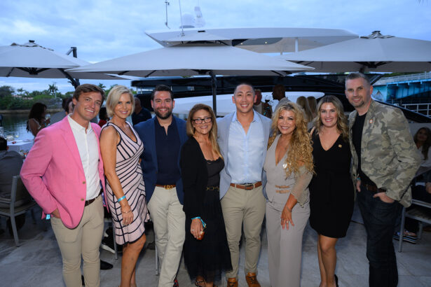 A group of eight people, dressed in semi-formal attire, pose and smile together outdoors near umbrellas and a docked yacht in the background. South Florida Business & Wealth