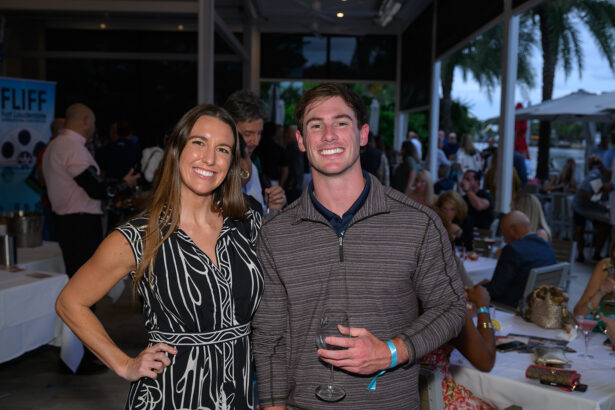 A smiling woman and man stand together at an outdoor event. The woman wears a black and white dress, and the man holds an empty wine glass. People are seated at tables in the background, with palm trees visible outside. South Florida Business & Wealth