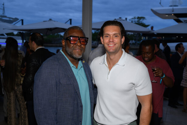 Two men posing and smiling at a waterfront event; one wears a blue blazer and large glasses, the other a white shirt. People and yachts are visible in the background, with umbrellas overhead. South Florida Business & Wealth