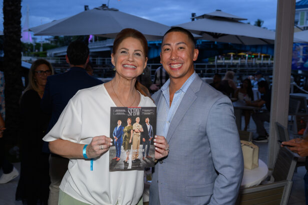 A smiling woman holding a SFBW magazine stands next to a smiling man in a light gray suit at an outdoor event with people, tables, and umbrellas in the background. South Florida Business & Wealth