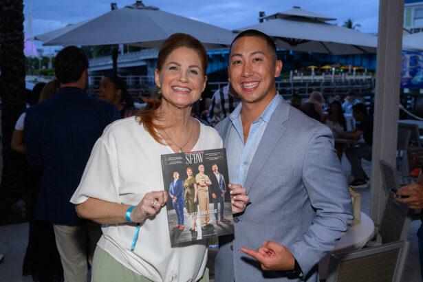 A smiling woman and man pose together outdoors at an event. The woman holds a magazine featuring four people on the cover, while the man points at it. Other attendees and patio umbrellas are visible in the background. South Florida Business & Wealth