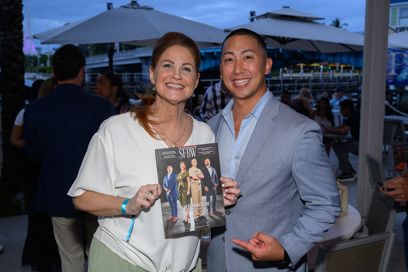 A smiling woman and man pose at an outdoor event; the woman holds up a "SFBW" magazine featuring business professionals on the cover, while the man points at the magazine. People and umbrellas are visible in the background. South Florida Business & Wealth