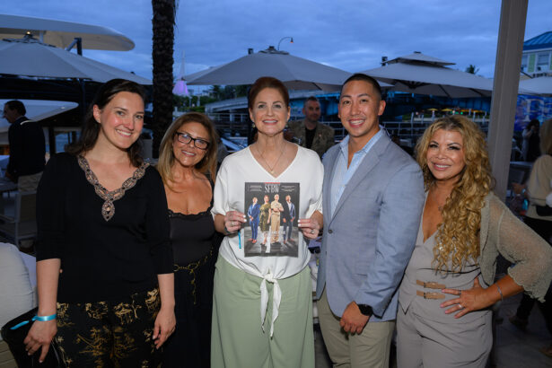 Five people stand together outdoors at a marina, smiling for the camera. The person in the center holds a magazine featuring a group photo on its cover. Umbrellas and yachts are visible in the background. South Florida Business & Wealth