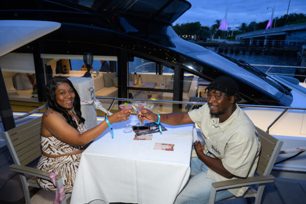 A smiling couple sits at a table on a boat, toasting with drinks. The woman wears a patterned dress, and the man a light shirt and cap. Evening sky and water are visible in the background. South Florida Business & Wealth