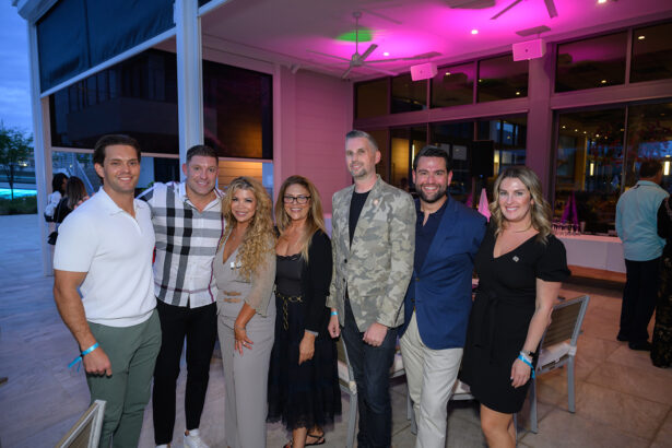 Seven adults stand together smiling at an outdoor evening event, dressed in semi-formal attire. Behind them are tables with white tablecloths and purple lighting, suggesting a social or celebratory gathering. South Florida Business & Wealth