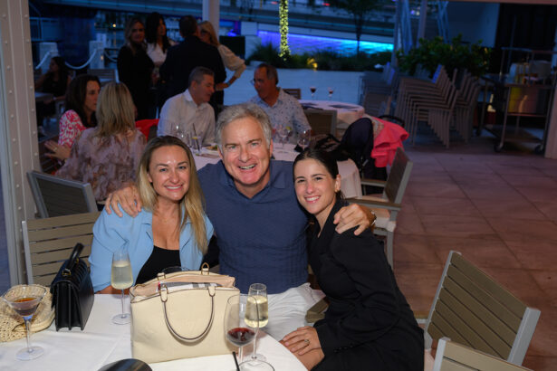 Three people sit closely together at an outdoor restaurant table, smiling at the camera. Wine glasses and a purse are on the table, with other diners and a lit pool area visible in the background. South Florida Business & Wealth