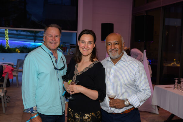 Three adults pose together at an indoor-outdoor event, smiling at the camera. Two men stand on either side of a woman holding a drink, while the men each hold wine glasses. Tables and lights are visible in the background. South Florida Business & Wealth