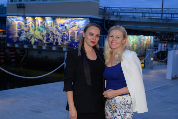 Two women pose and smile outdoors in the evening. One wears a black suit and red lipstick, the other a white jacket over a blue top with printed pants. A colorful mural and a bridge are visible in the background. South Florida Business & Wealth