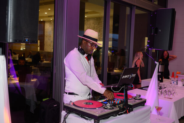 A DJ wearing a white suit, hat, and headphones is mixing music on turntables at an indoor event, with people socializing and dancing in the background. South Florida Business & Wealth