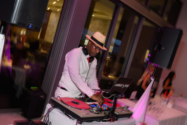 A DJ wearing a white shirt, red tie, and a straw hat mixes music on turntables at an indoor event, surrounded by party guests and ambient lighting. South Florida Business & Wealth