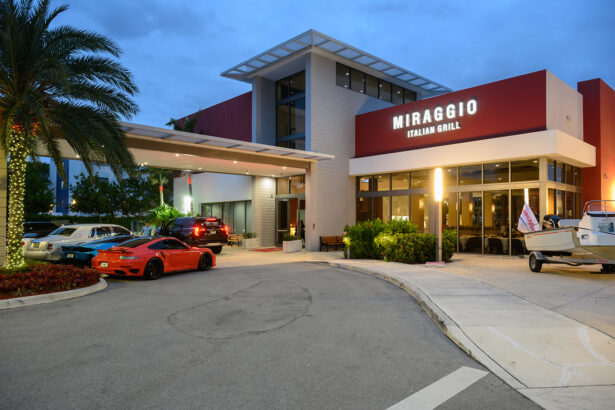 The exterior of Miraggio Italian Grill at dusk, with lights on, a red sports car and other vehicles parked out front, a palm tree with string lights, and a small boat on a trailer near the entrance. South Florida Business & Wealth