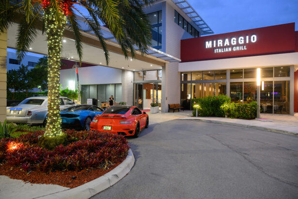 Three luxury cars parked near the entrance of Miraggio Italian Grill at dusk, with palm trees wrapped in string lights and modern building architecture visible. South Florida Business & Wealth