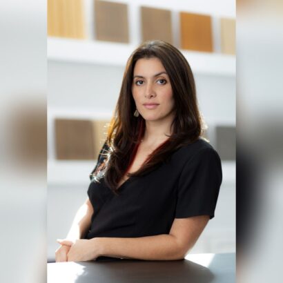 A woman with long dark hair wearing a black top sits at a table, looking confidently at the camera. The background features blurred rectangular color samples on the wall. South Florida Business & Wealth
