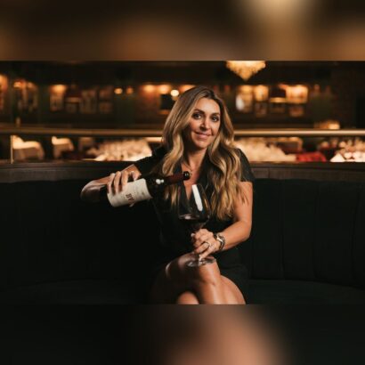 A woman with long hair sits on a dark sofa in a dimly lit restaurant, smiling as she pours red wine from a bottle into a glass, with elegant decor visible in the background. South Florida Business & Wealth