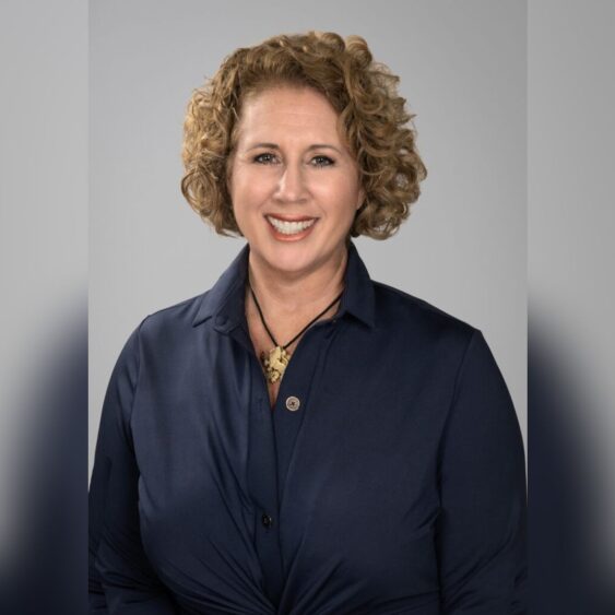 A woman with curly light brown hair, wearing a dark blue collared shirt and a gold pendant necklace, smiles at the camera against a plain gray background. South Florida Business & Wealth