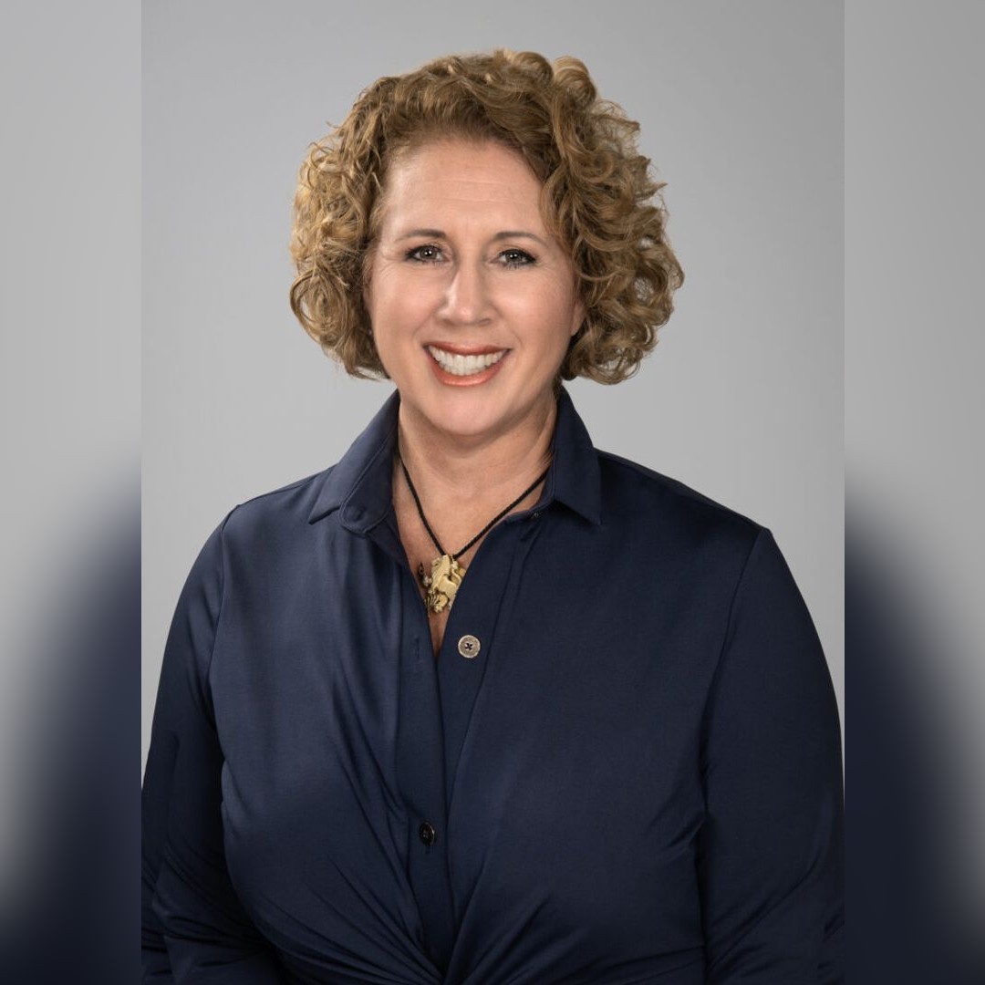 A woman with curly light brown hair, wearing a dark blue collared shirt and a gold pendant necklace, smiles at the camera against a plain gray background. South Florida Business & Wealth