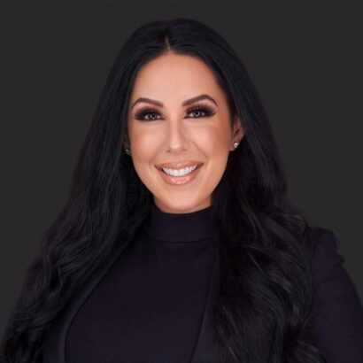 A woman with long, wavy dark hair wearing a black blazer and top smiles at the camera against a plain dark background. South Florida Business & Wealth