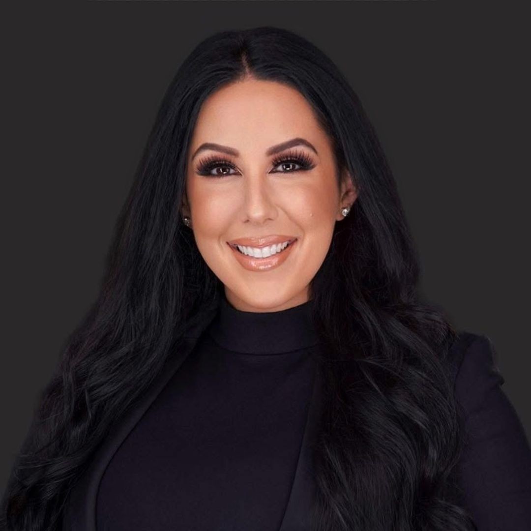 A woman with long, wavy dark hair wearing a black blazer and top smiles at the camera against a plain dark background. South Florida Business & Wealth
