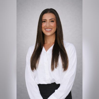 A woman with long straight brown hair, wearing a white button-up blouse and black pants, smiles at the camera against a plain light gray background. South Florida Business & Wealth