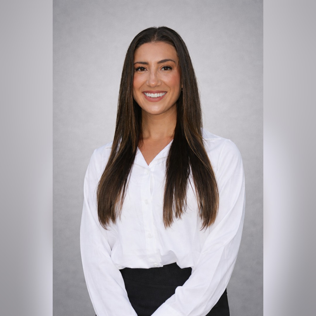 A woman with long straight brown hair, wearing a white button-up blouse and black pants, smiles at the camera against a plain light gray background. South Florida Business & Wealth
