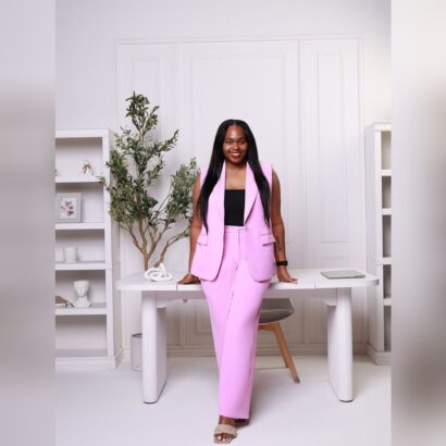 A woman wearing a pink pantsuit and black top smiles while leaning against a white desk in a modern, minimal office with shelves, plants, and decor in the background. South Florida Business & Wealth