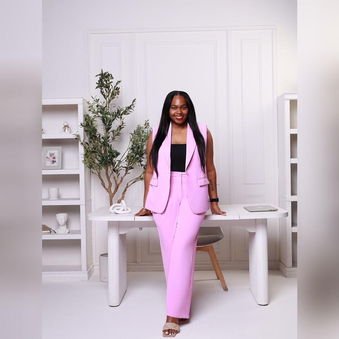 A woman wearing a pink pantsuit and black top smiles while leaning against a white desk in a modern, minimal office with shelves, plants, and decor in the background. South Florida Business & Wealth
