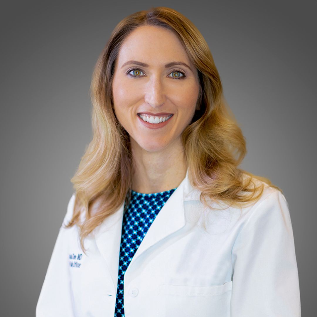 A woman with long, wavy blonde hair, wearing a white medical coat over a blue patterned top, smiles at the camera against a plain gray background. South Florida Business & Wealth