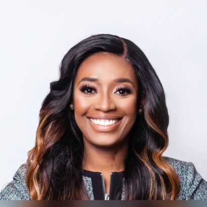 A woman with long, wavy dark hair and caramel highlights smiles warmly at the camera. She is wearing a textured, patterned jacket and has a light background behind her. South Florida Business & Wealth