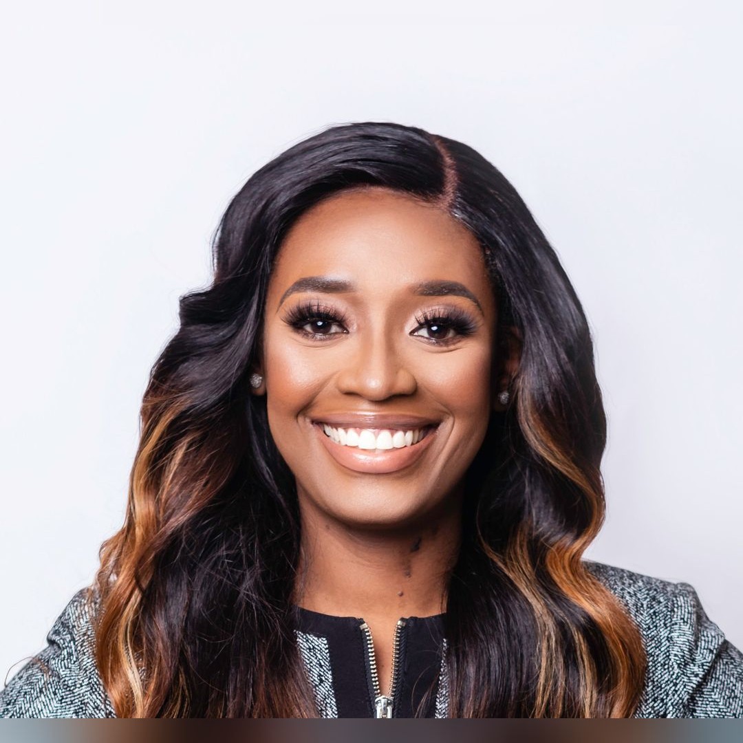 A woman with long, wavy dark hair and caramel highlights smiles warmly at the camera. She is wearing a textured, patterned jacket and has a light background behind her. South Florida Business & Wealth
