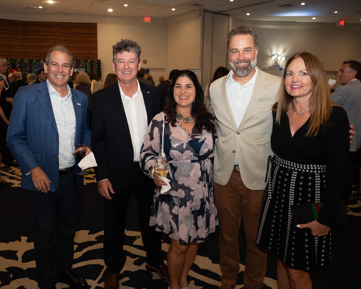 Five adults, dressed in semi-formal attire, stand together smiling at an indoor event. The group includes three men in jackets and two women in dresses, with other guests visible in the background. South Florida Business & Wealth