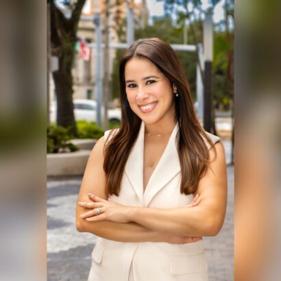 A woman with long brown hair, wearing a white sleeveless blazer, stands outdoors with arms crossed and smiles at the camera. Trees and buildings are visible in the blurred background. South Florida Business & Wealth