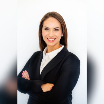 A woman with straight brown hair wearing a black blazer and white shirt stands smiling confidently with her arms crossed against a plain white background. South Florida Business & Wealth
