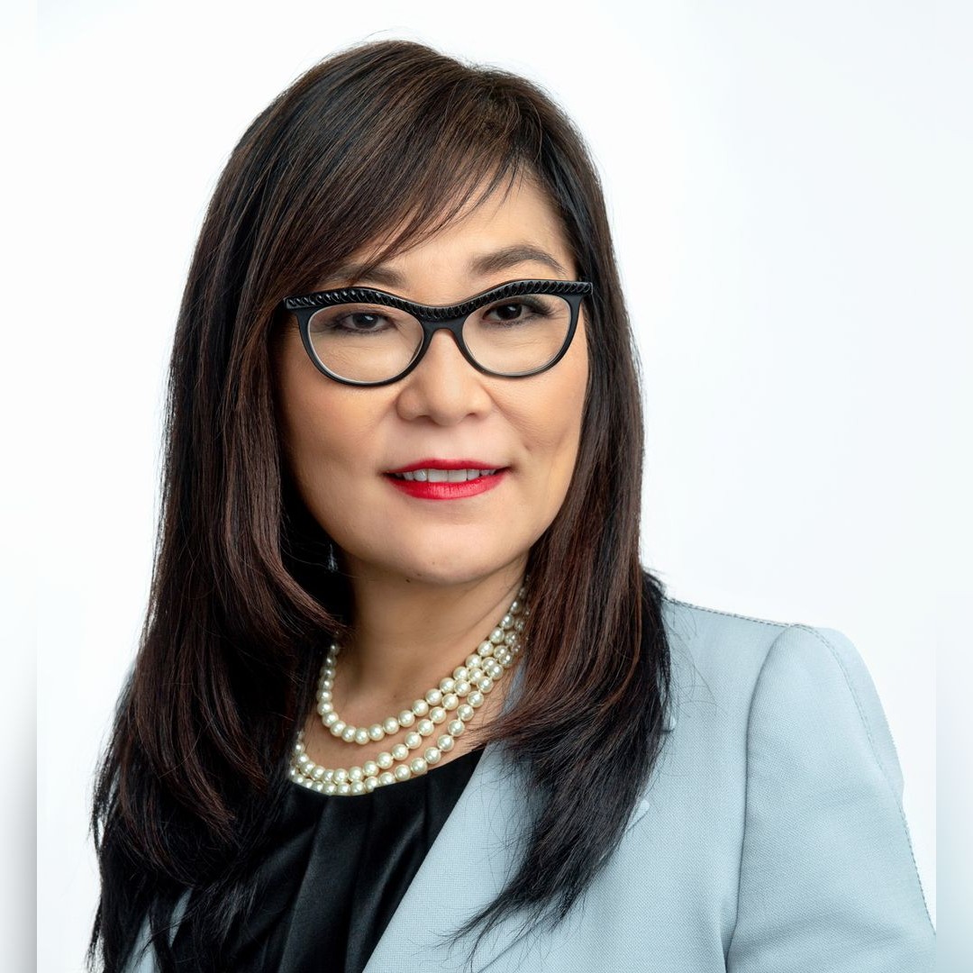 A woman with long dark hair wearing black cat-eye glasses, a light gray blazer over a black top, red lipstick, and layered pearl necklaces, posed against a plain white background. South Florida Business & Wealth