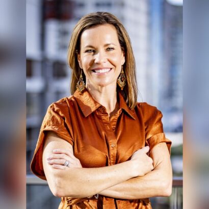 A woman with straight brown hair wearing a shiny orange blouse and dangling earrings stands outdoors with her arms crossed, smiling confidently. The background shows city buildings out of focus. South Florida Business & Wealth