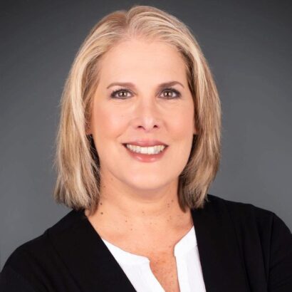 A woman with shoulder-length blonde hair smiles at the camera. She is wearing a black blazer over a white blouse and poses in front of a plain dark gray background. South Florida Business & Wealth