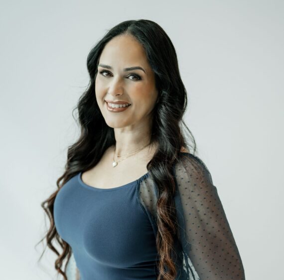 A woman with long, wavy dark hair smiles at the camera. She is wearing a sheer-sleeved, navy blue top and a delicate necklace. The background is plain and light-colored. South Florida Business & Wealth