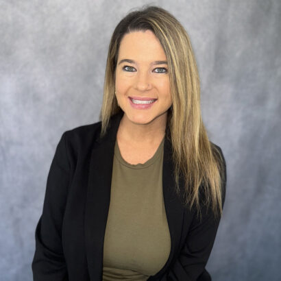 A woman with straight blonde hair wearing a black blazer over an olive green top smiles at the camera, sitting in front of a gray background. South Florida Business & Wealth