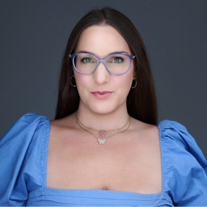 A woman with long brown hair wearing blue glasses, a blue square-neck top with puffed sleeves, gold hoop earrings, and layered necklaces, poses against a dark gray background. South Florida Business & Wealth