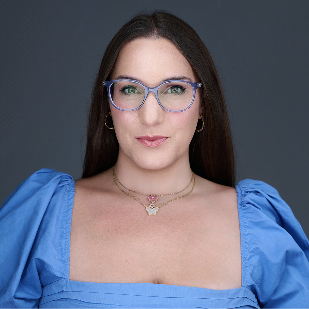A woman with long brown hair wearing blue glasses, a blue square-neck top with puffed sleeves, gold hoop earrings, and layered necklaces, poses against a dark gray background. South Florida Business & Wealth
