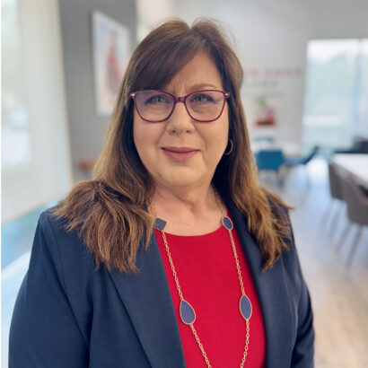 A woman with long brown hair wearing glasses, a navy blazer, and a red top stands in a modern office space, smiling at the camera. She wears a gold necklace with blue pendants. The background is softly blurred. South Florida Business & Wealth