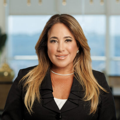 A woman with long light brown hair, wearing a black blazer over a white top and a pearl necklace, smiles at the camera in a modern office setting with blurred windows in the background. South Florida Business & Wealth